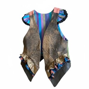 May include: A brown leather vest with a distressed finish. The vest has a striped fabric lining in purple, blue, and green. Gold trim and floral patterned fabric adorn the bottom edges. The sleeves have ruffled edges with black polka dots.