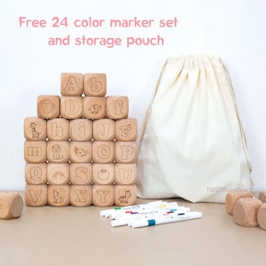 May include: A set of light-colored wooden alphabet blocks featuring engraved letters and animal illustrations, accompanied by a beige drawstring storage pouch and a collection of colorful markers. The text Free 24 color marker set and storage pouch is visible.