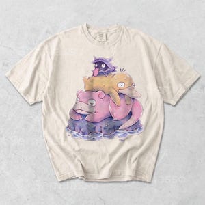 May include: Cream-colored t-shirt featuring a watercolor illustration of three cartoon creatures stacked on top of each other. The creatures are pink, yellow, and purple, with a blue and purple base. The shirt has a crew neck and short sleeves.