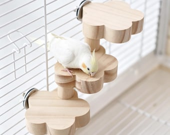 May include: A light-coloured wooden bird perch set featuring three flower-shaped platforms attached to a white wire cage. A small white cockatiel with yellow head feathers stands on the middle platform, pecking at the wood surface.