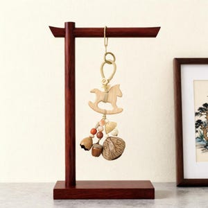 May include: A dark wood T-shaped display stand holding a hanging ornament. The ornament features a light wood rocking horse cutout, a beige cord loop, and a cluster of natural elements including small wooden beads, an acorn, and a large seed pod.