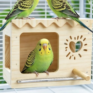 Op de afbeelding: Een lichtgekleurd houten vogelhuisje met een geschulpte dakrand, een grote vierkante opening en een hartvormige uitsparing omringd door stralende lijnen. Een kleine houten zitstok steekt aan de voorzijde uit en drie groen-gele parkieten zijn zichtbaar.