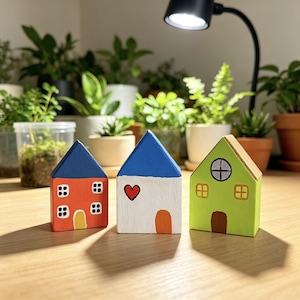 Puede incluir: Tres pequeñas figuras de casas de madera pintadas en colores brillantes. La primera es naranja con techo azul, la segunda es blanca con techo azul y un corazón rojo, y la tercera es verde lima con una puerta marrón y una ventana circular.