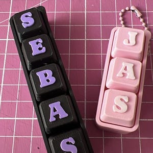 May include: Two custom name keychains. One black keychain spells out "SEBAS" in purple letters. The other is pink and spells out "JAS" in white letters. Both are on a pink grid background.