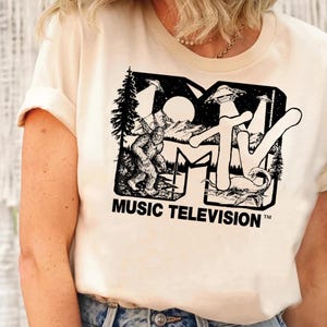 May include: A cream-colored short-sleeve t-shirt featuring a black graphic print of the MTV logo. Inside the logo letters are illustrations of Bigfoot, a Loch Ness monster, mountains, trees, and flying saucers. The text MUSIC TELEVISION is below.