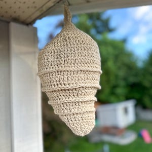 Op de afbeelding: Een beige, gehaakte hangdecoratie, die lijkt op een traan of een cocon. Het handgemaakte item is opgehangen aan een haak, met een spiraalvormig ontwerp met zichtbare steekpatronen. De achtergrond is wazig, met hints van groen en een blauwe lucht.