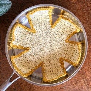 Könnte beinhalten: Ein cremefarbener, gehäkelter Topflappen mit gelbem Rand, der in einer Edelstahlpfanne liegt. Der Topflappen hat ein Sonnenmuster. Die Pfanne steht auf einer Holzoberfläche.