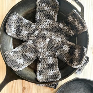 Può includere: Una presina fatta a mano all'uncinetto in una padella di ghisa. La presina è un mix di filato grigio, nero e bianco, con un centro circolare e sei sezioni estese. La padella è nera e poggia su una superficie di legno chiaro.