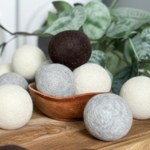 Peut inclure: Un assortiment de boules de séchage en laine feutrée de différentes couleurs, dont blanc, gris et marron. Les boules sont disposées dans un bol en bois et sur une surface en bois, avec un feuillage vert en arrière-plan. Solution de séchage durable.