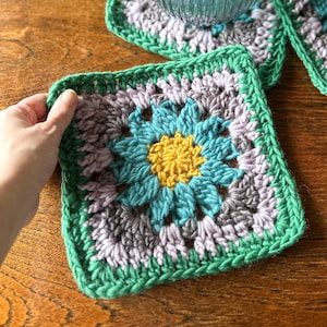 May include: Handmade crochet coasters in a floral design. The coasters feature a central yellow flower surrounded by teal petals, grey and lavender accents, and a green border. The square coasters are approximately 13 cm by 13 cm.