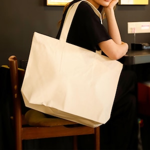 May include: A large, off-white canvas tote bag with matching straps. The bag is rectangular and appears to be empty. The tote bag is being held by a person wearing a black shirt.