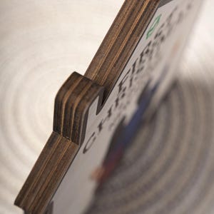 May include: A house-shaped wooden plaque with visible layered plywood edges. The front surface is white and features a partial graphic print with the text "CH-FIB" and a small green icon. The object is shown from an angled side perspective.