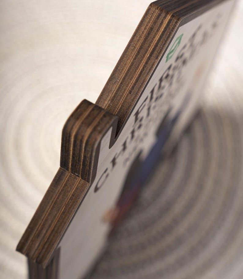 May include: A house-shaped wooden plaque with visible layered plywood edges. The front surface is white and features a partial graphic print with the text "CH-FIB" and a small green icon. The object is shown from an angled side perspective.