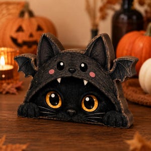 May include: A black cat figurine wearing a bat-themed hood with small wings, pointed ears, and pink cheeks. The cat has large, bright orange eyes, visible white fangs, and two front paws resting at the base of the dark, textured wooden structure.