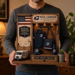 May include: A wooden display featuring a miniature mail truck, a personalized plaque for Michael Johnson, a small USPS jacket, a shoulder bag, a cap, and a water bottle. The unit includes text reading Mail Carrier, Delivering More Than Mail, They Deliver Hope, and Service, Integrity, Connection.