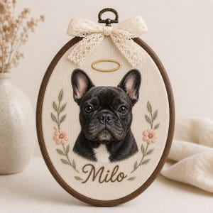 Puede incluir: Un bastidor de bordado ovalado con el retrato de un bulldog francés negro con una mancha blanca en el pecho. Sobre el perro hay un halo dorado bordado. Debajo, el nombre Milo está bordado en marrón. Incluye ramas florales y un lazo de encaje.