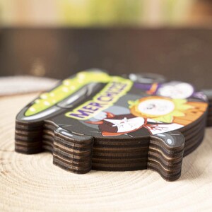 May include: A layered wooden cutout shaped like a cauldron with a black exterior and a bright green liquid interior. The surface features a cartoon illustration of a cat and a pumpkin with the text MER CHIZZA printed in purple letters.