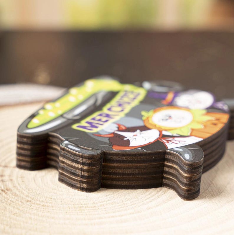 May include: A layered wooden cutout shaped like a cauldron with a black exterior and a bright green liquid interior. The surface features a cartoon illustration of a cat and a pumpkin with the text MER CHIZZA printed in purple letters.
