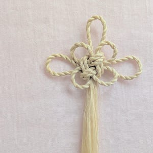 以下が含まれることがあります： ねじれたロープで作られた、花に似たクリーム色の装飾的な結び目。 結び目は、ループ状の花びらと長いタッセルが付いた中央の織り部分があります。 背景は柔らかなピンク色の生地です。