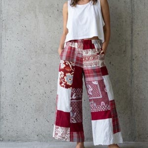 Könnte beinhalten: Weite Patchworkhose in Rot, Weiß und Burgunder. Die Hose hat eine Vielzahl von gemusterten Stoffquadraten, darunter Blumen- und Karomuster. Das Model trägt ein weißes Tanktop. Die Hose hat Taschen.