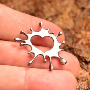 May include: A silver-colored pendant in the shape of a splash with a heart-shaped cutout in the center. The pendant has a textured, liquid-like design with droplet details. The jewelry is held in a hand against a blurred, natural background.