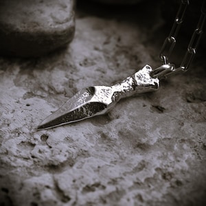 Peut inclure: Un collier pendentif argenté avec un charme en forme de dague. Le charme a un aspect texturé et martelé et une pointe. La chaîne est de style maillon simple. Le collier est présenté sur une surface grise texturée.