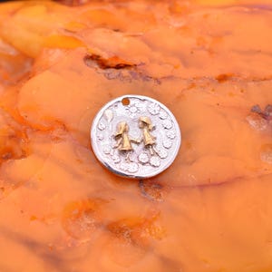 Könnte beinhalten: Ein silberfarbener Anhänger mit detailliertem Design. Der Anhänger zeigt zwei kleine Figuren, die sich an den Händen halten, umgeben von floralen und Sonnenmotiven. Die Figuren haben goldfarbene Kleider und Hüte. Der Anhänger hat oben ein kleines Loch für eine Kette.