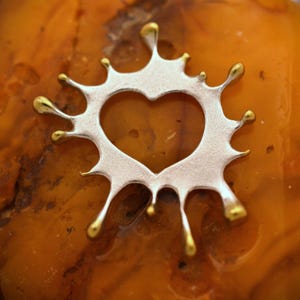 May include: A silver-colored pendant with a heart-shaped cutout, accented with gold-colored droplets. The pendant has a unique splash design. The background is a textured, orange-brown surface.