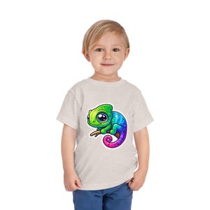 May include: A light beige toddler t-shirt featuring a colorful cartoon chameleon perched on a branch. The chameleon has green, blue, and purple hues. The t-shirt is made of a soft, comfortable material.