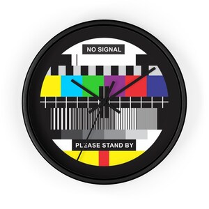 Puede incluir: Un reloj de pared redondo negro con un diseño de patrón de prueba colorido. La esfera del reloj incluye el texto "NO SIGNAL" en la parte superior y "PLEASE STAND BY" en la parte inferior. Las manecillas del reloj son negras.