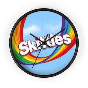 May include: Round wall clock with a black frame. The clock face features the word "Skittles" in white with a red outline, set against a blue sky background with a rainbow.