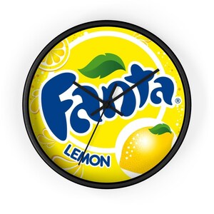 May include: Round wall clock with a black frame. The clock face is yellow with the Fanta logo in blue, a lemon graphic, and the word "LEMON". The clock hands are black.