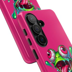 May include: Bright pink phone case with a cartoon frog design. The frog has large red eyes and a wide open mouth, with green and pink detailing. The case includes a camera and side buttons.