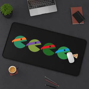 May include: A black rectangular desk mat featuring a graphic design of four green circular characters with masks in orange, purple, red, and blue. The characters are arranged in a row, with the last one holding a slice of pepperoni pizza.
