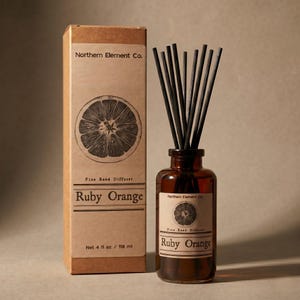 May include: A brown glass bottle diffuser with black reeds and a matching box. The box and label on the bottle read "Ruby Orange" and "Northern Element Co." The box also states "Net 4 fl oz / 118 ml". The design features a cross-section of an orange.