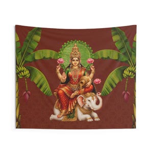 May include: A rectangular wall tapestry featuring a central illustration of the Hindu goddess Lakshmi seated on a white elephant. She wears ornate gold jewelry and a red and green sari, holding pink lotus flowers and a pot pouring gold coins.