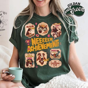 May include: Forest green t-shirt with multiple cartoon graphics. The graphics include a cat with a crown, a dog at a computer, a goat, a dinosaur, and text that reads "NEEEEEW ACHIEVEMENT!" and "The Desperado Club".