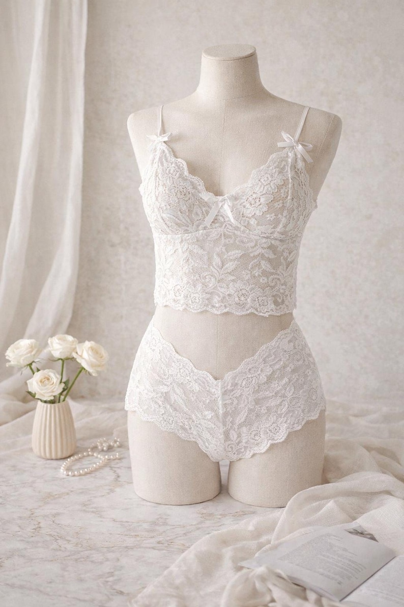 May include: A white lace lingerie set on a mannequin. The set features a bralette with bow details and matching briefs. The delicate floral lace design is visible. A vase of white roses and a string of pearls are in the background.