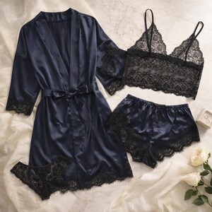 May include: A navy blue satin lingerie set comprising a robe, bralette, shorts, and underwear. The robe has a tie closure and lace trim on the sleeves. The bralette and underwear are black lace. The shorts have lace trim. A white rose and a ring box are also in the image.