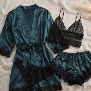 May include: A teal satin pyjama set with black lace trim. The set includes a robe with a tie, a bralette, and shorts. The bralette and shorts have black lace detailing. The set is displayed on a white surface.