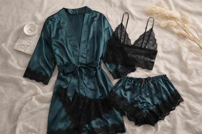 May include: A teal satin pyjama set with black lace trim. The set includes a robe with a tie, a bralette, and shorts. The bralette and shorts have black lace detailing. The set is displayed on a white surface.
