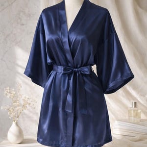 May include: A navy blue satin robe with a matching tie belt, displayed on a mannequin. The robe has wide sleeves and a V-neck design. The fabric has a subtle sheen, and the robe is approximately knee-length.