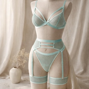 May include: A light blue lingerie set displayed on a mannequin. The set includes a bra, high-waisted briefs, and a garter belt with suspenders. The bra and briefs have a textured pattern. The garter belt has a delicate design.