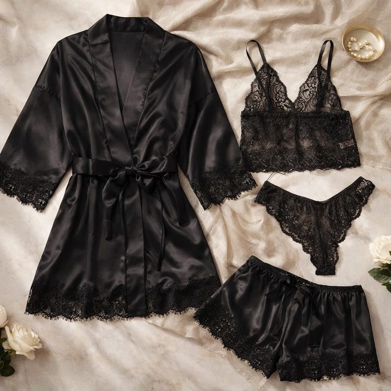 May include: A black satin lingerie set comprising a robe with lace trim, a lace bralette, a pair of lace thong underwear, and satin shorts with lace trim. The set is arranged on a neutral fabric.