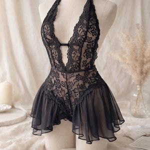 Puede incluir: Lencería de encaje negro con cuello halter y escote en V pronunciado. El body presenta un patrón de encaje floral y una falda con volantes transparente. La prenda se exhibe en un maniquí.