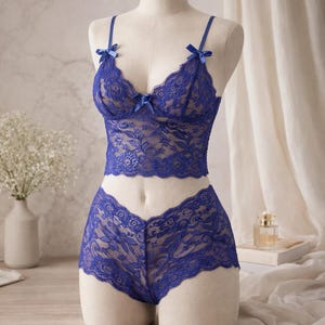 May include: A blue lace lingerie set displayed on a mannequin. The set includes a bralette with bow accents and a matching low-rise bottom. The delicate floral lace design is visible throughout the set. The lingerie is displayed on a neutral background.