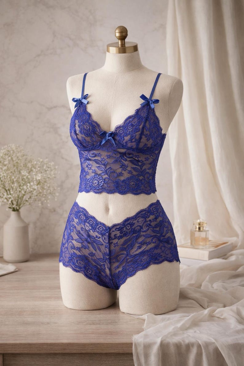 May include: A blue lace lingerie set on a mannequin. The set features a bralette with bow details and matching low-rise briefs. The intricate floral lace pattern is visible. The lingerie is displayed against a neutral backdrop.