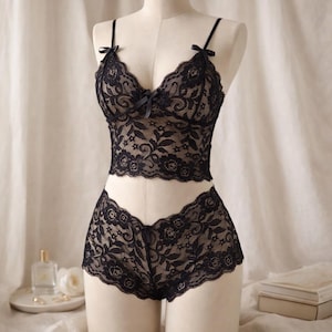 May include: Black lace lingerie set on a mannequin. The set features a camisole top with thin straps and a bow, and matching shorts. The delicate floral pattern is visible throughout the set.