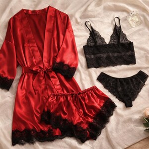 May include: A red satin robe with black lace trim, a matching shorts set, a black lace bralette, and a black lace thong. The lingerie set is displayed on a white sheet with a perfume bottle and a bouquet of flowers.