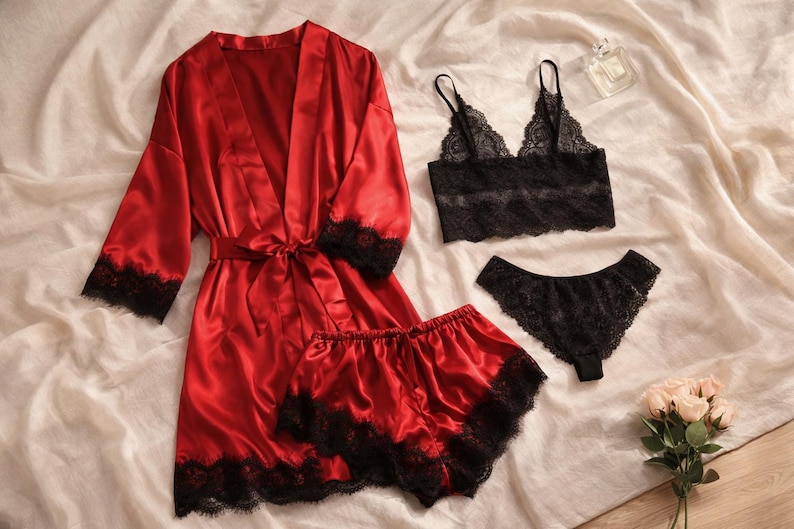 May include: A red satin robe with black lace trim, a matching shorts set, a black lace bralette, and a black lace thong. The lingerie set is displayed on a white sheet with a perfume bottle and a bouquet of flowers.